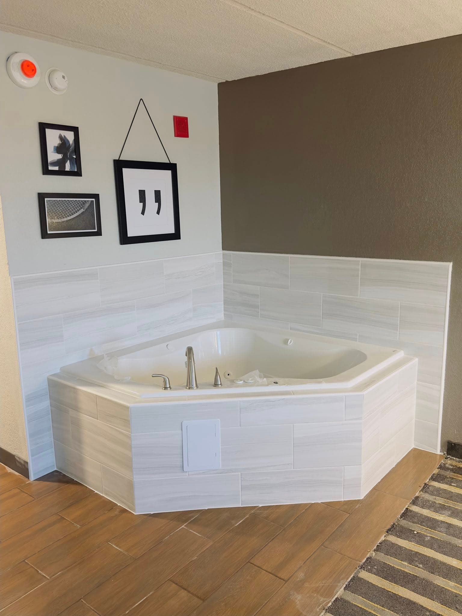 White corner whirlpool tub with light wood-patterned tiles and framed artwork on a gray wall.